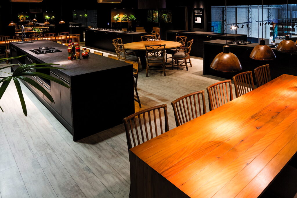 Mesa longa de madeira maciça do Amazônia Gourmet sob as luminárias de cobre, ao lado das bancadas de cocção