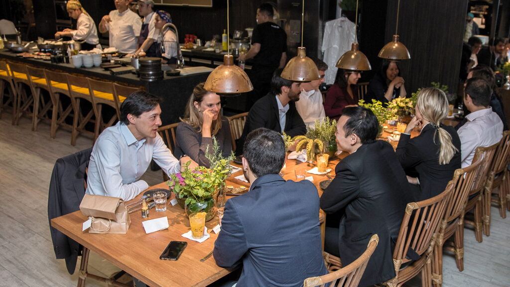Convidados conversam na mesa longa do Amazônia Gourmet com a cozinha em operação ao fundo