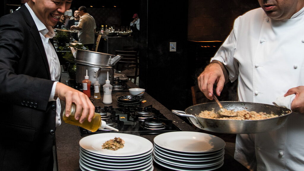 Participante de terno finaliza o prato na bancada ao lado do chef da casa durante team building gastronômico